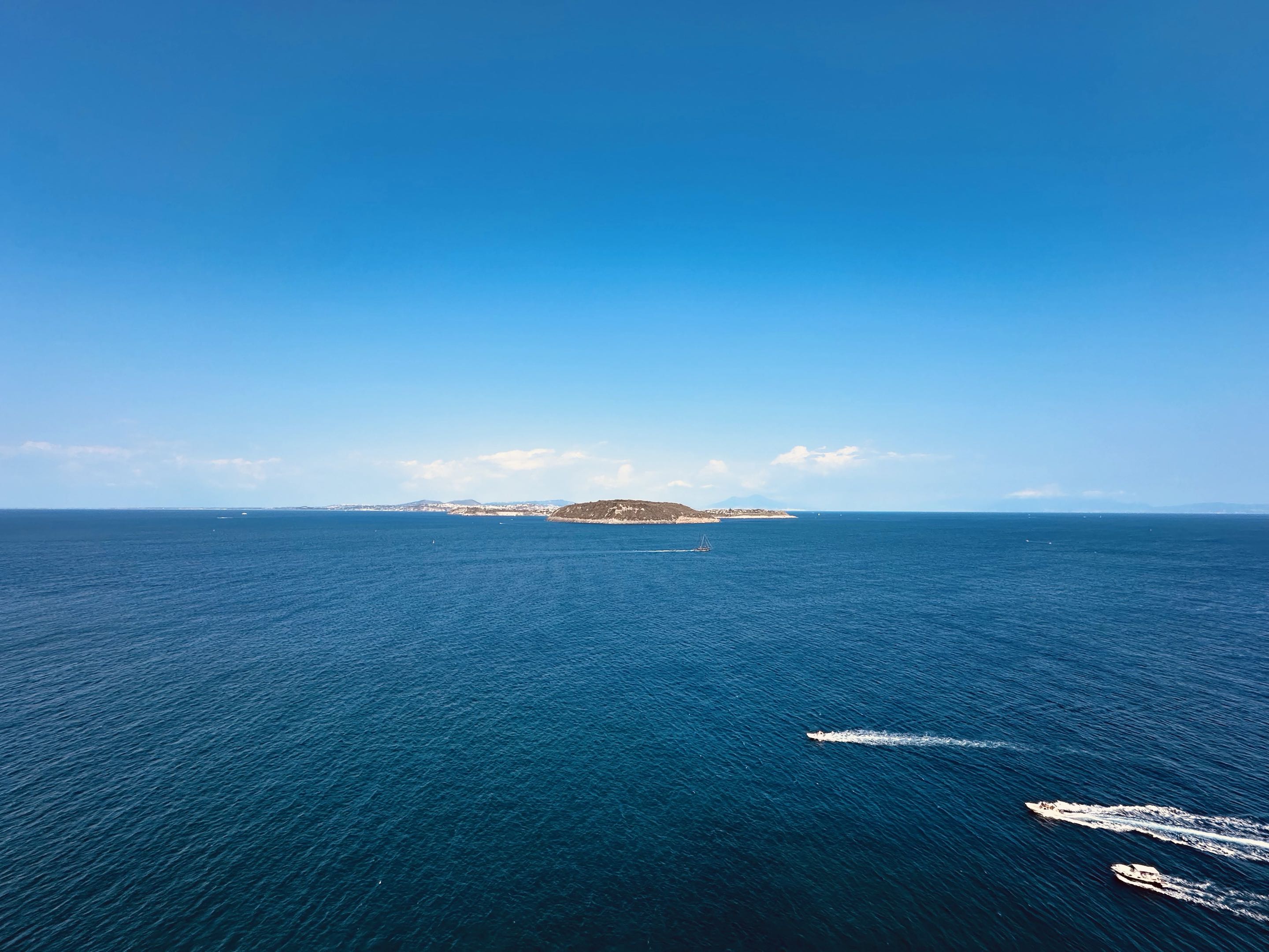 Motorboats leave white wakes across deep blue water off Ischia, with low islands on the horizon.