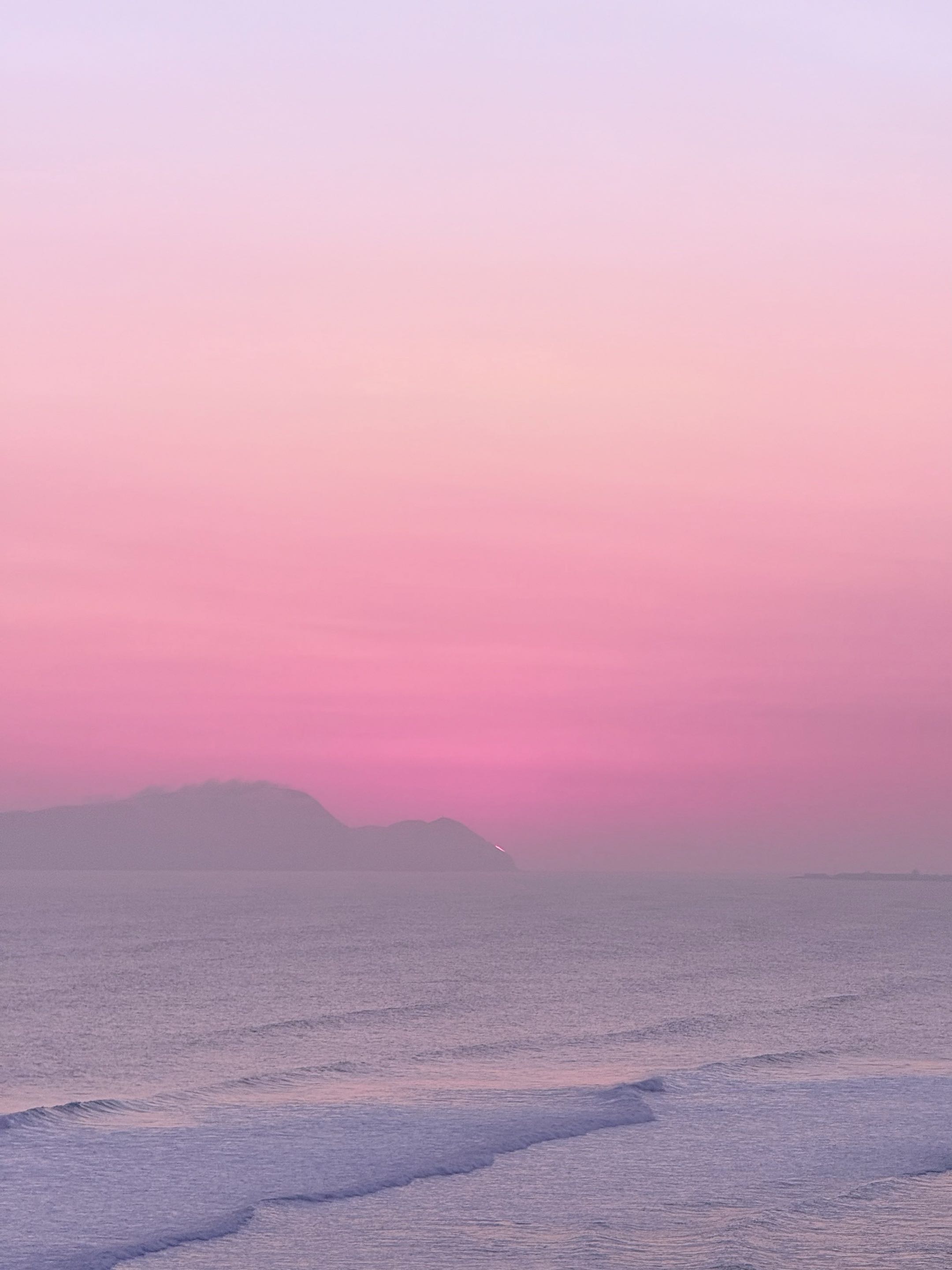 Soft pink sunset over the Pacific, with waves and a hazy island silhouette off Lima.
